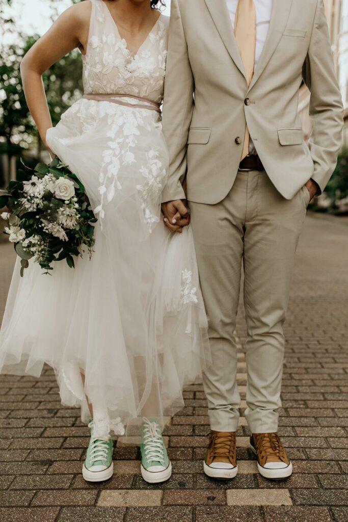 Mann in grauer Anzugjacke und Frau in weißem Brautkleid mit weißem Blumenstrauß und Converse Schuhen